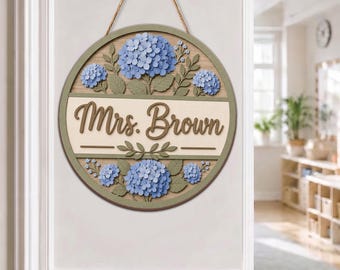 May include: A circular wooden wall sign featuring a light wood grain background, a central cream-coloured horizontal band with the text "Mrs. Brown" in brown script, and layered blue hydrangea floral cutouts with green leaves, suspended by a twine rope.