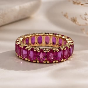 Peut inclure: Une bague alliance ornée d'une rangée continue de rubis rose vif taille ovale, sertis dans une monture en métal doré jaune poli. Les pierres précieuses sont disposées côte à côte, formant un design circulaire avec une finition métallique.
