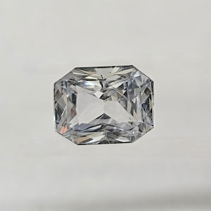May include: A clear, rectangular-cut gemstone with multiple facets, reflecting light. The gem is set against a white background, showcasing its clarity and brilliance. The cut is symmetrical, with a flat top and angled sides.