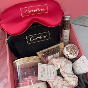 Op de afbeelding: Een roze geschenkdoos met het label "Scarlet Elegance Box" gevuld met schoonheidsproducten. Inclusief een rood satijnen oogmasker, een zwarte cosmetische tas met "Caroline" in goud, een kaars, badproducten en een bloemen scrunchie. Van "Beauty Mix Shop".