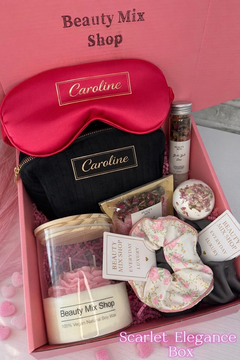 Op de afbeelding: Een roze geschenkdoos met het label "Scarlet Elegance Box" gevuld met schoonheidsproducten. Inclusief een rood satijnen oogmasker, een zwarte cosmetische tas met "Caroline" in goud, een kaars, badproducten en een bloemen scrunchie. Van "Beauty Mix Shop".