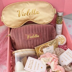 Op de afbeelding: Een roze geschenkdoos met het label "Champagne Glow Box" gevuld met schoonheidsproducten. Inclusief een gouden oogmasker en een mauve cosmetische tas, beide met "Violetta" in gouden letters. Bevat ook een kaars, badzout en andere items.