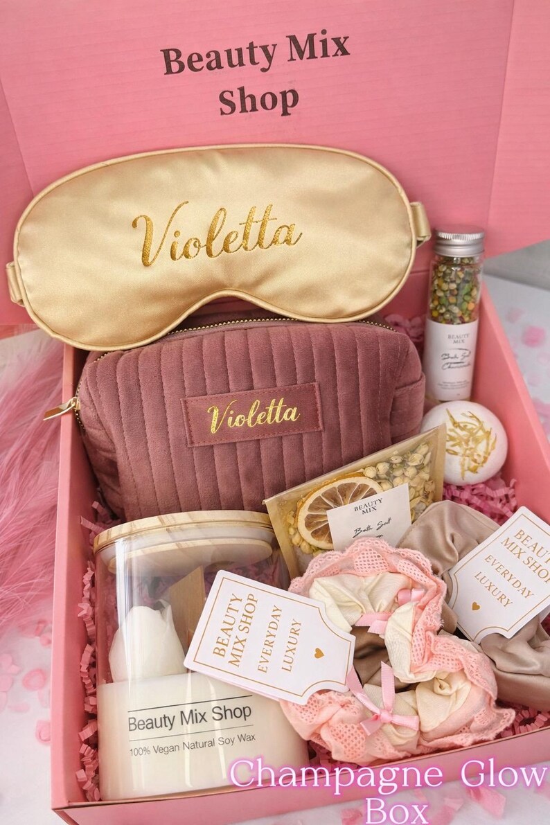 Op de afbeelding: Een roze geschenkdoos met het label "Champagne Glow Box" gevuld met schoonheidsproducten. Inclusief een gouden oogmasker en een mauve cosmetische tas, beide met "Violetta" in gouden letters. Bevat ook een kaars, badzout en andere items.