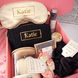 Op de afbeelding: Een roze geschenkdoos met de tekst "Midnight Luxe Box" gevuld met schoonheidsproducten. Items zijn een slaapmasker en een cosmetische tas, beide met "Katie" in gouden letters, een kaars, badzout, een badbruisbal, scrunchies en een theezakje.