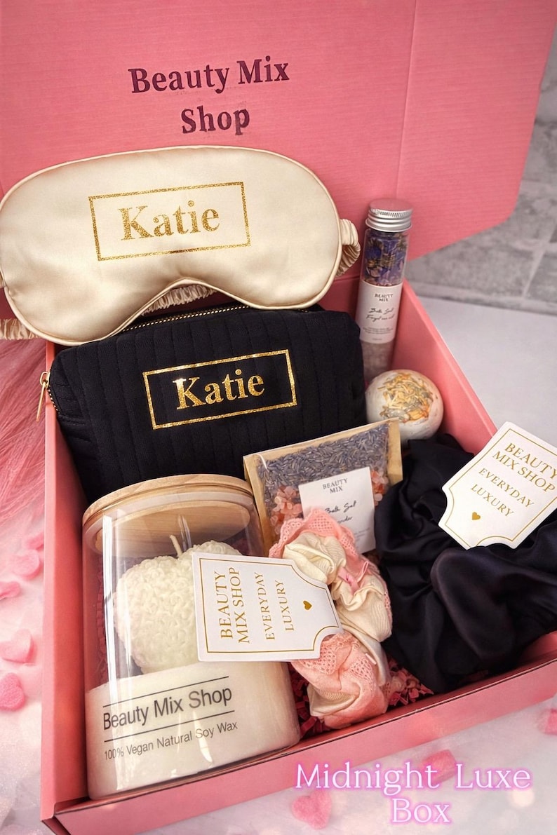 Op de afbeelding: Een roze geschenkdoos met de tekst "Midnight Luxe Box" gevuld met schoonheidsproducten. Items zijn een slaapmasker en een cosmetische tas, beide met "Katie" in gouden letters, een kaars, badzout, een badbruisbal, scrunchies en een theezakje.