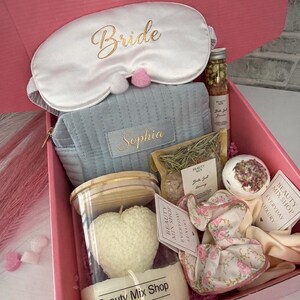 Op de afbeelding: Een roze geschenkdoos met het opschrift "Bridal Bliss Box" gevuld met zelfzorgartikelen. Inclusief een wit oogmasker met "Bride", een blauw etui met "Sophia", een hartvormige kaars, badproducten en bloemaccessoires.