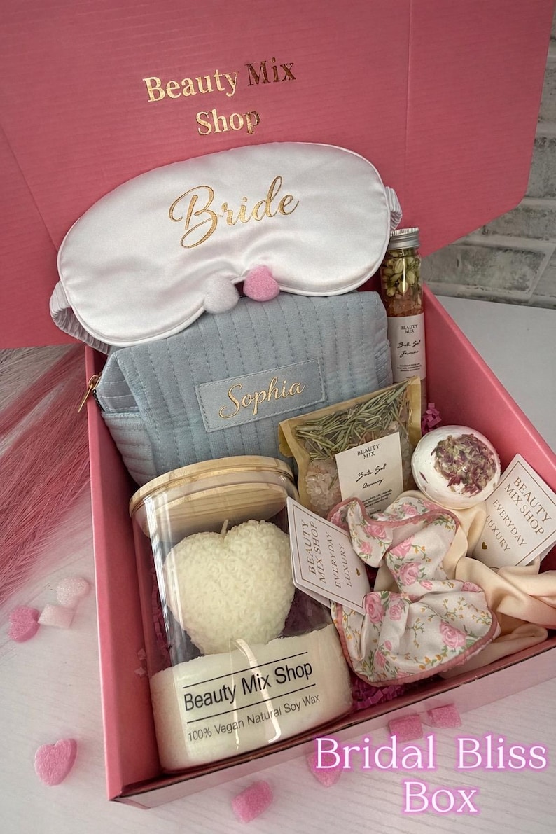 Op de afbeelding: Een roze geschenkdoos met het opschrift "Bridal Bliss Box" gevuld met zelfzorgartikelen. Inclusief een wit oogmasker met "Bride", een blauw etui met "Sophia", een hartvormige kaars, badproducten en bloemaccessoires.