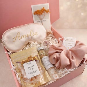 Op de afbeelding: Een roze geschenkdoos gevuld met zelfzorgartikelen. Inclusief een satijnen oogmasker met de naam "Amelia", badzout, een scrunchie en een kaart met gedroogde bloemen. De doos is gevuld met wit papier.