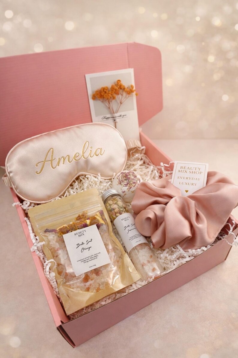 Op de afbeelding: Een roze geschenkdoos gevuld met zelfzorgartikelen. Inclusief een satijnen oogmasker met de naam "Amelia", badzout, een scrunchie en een kaart met gedroogde bloemen. De doos is gevuld met wit papier.