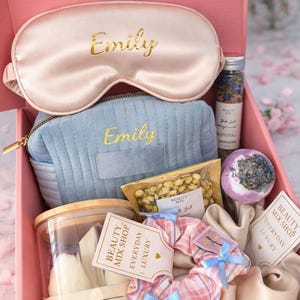 Op de afbeelding: Een roze geschenkdoos van "Beauty Mix Shop" met schoonheidsproducten. Het bevat een satijnen oogmasker en een cosmetische tas met de borduring "Emily", een kaars, een badbruisbal en andere items. De doos is gelabeld "Soft Cloud Box".