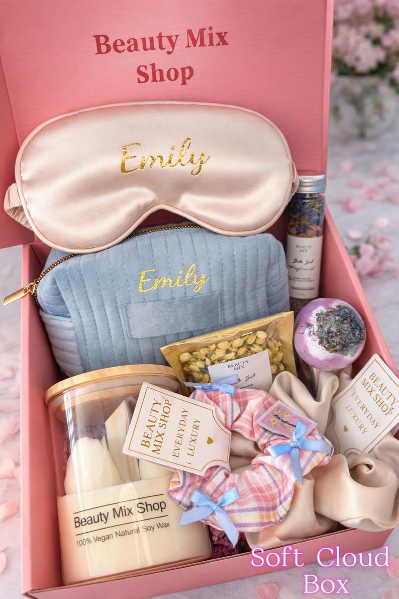 Op de afbeelding: Een roze geschenkdoos van "Beauty Mix Shop" met schoonheidsproducten. Het bevat een satijnen oogmasker en een cosmetische tas met de borduring "Emily", een kaars, een badbruisbal en andere items. De doos is gelabeld "Soft Cloud Box".