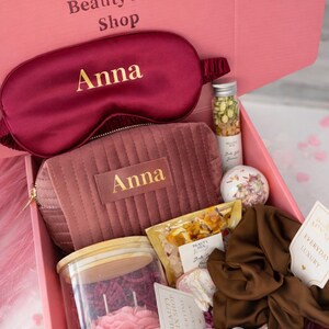 Op de afbeelding: Een roze geschenkdoos met het label "Velvet Rose Box" gevuld met schoonheidsproducten. Bevat een bordeauxkleurig oogmasker en een bijpassende cosmetische tas, beide gepersonaliseerd met "Anna" in gouden letters. Bevat ook een kaars, badzout en andere luxeartikelen.