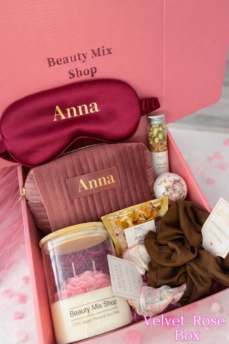 Op de afbeelding: Een roze geschenkdoos met het label "Velvet Rose Box" gevuld met schoonheidsproducten. Bevat een bordeauxkleurig oogmasker en een bijpassende cosmetische tas, beide gepersonaliseerd met "Anna" in gouden letters. Bevat ook een kaars, badzout en andere luxeartikelen.