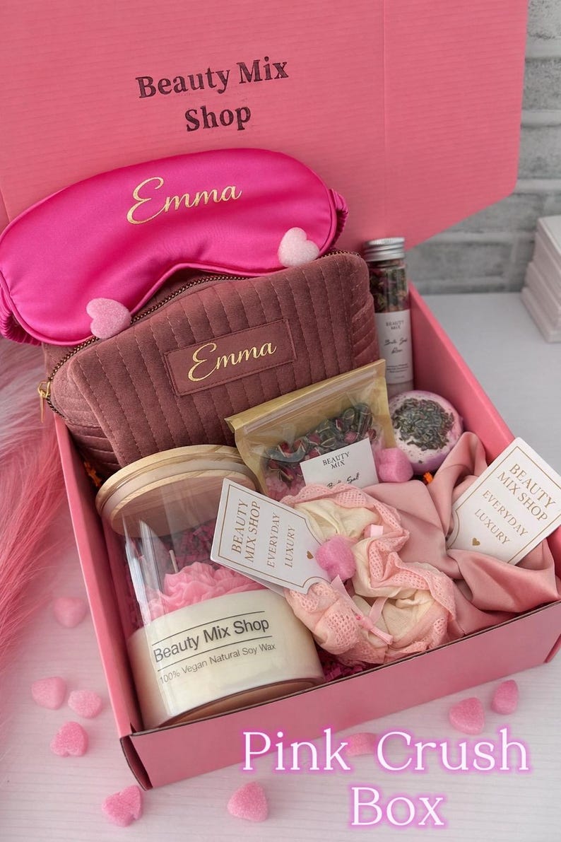 Op de afbeelding: Een roze geschenkdoos met het label "Pink Crush Box" gevuld met schoonheidsproducten. Inclusief een slaapmasker en een cosmetische tas, beide met "Emma" erop gedrukt. Bevat ook een kaars, badbruisbal en andere items.