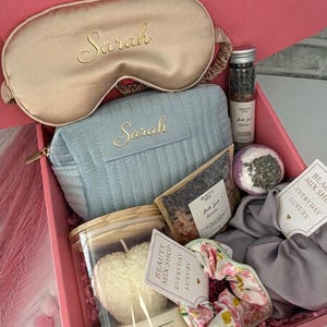 Op de afbeelding: Een roze geschenkdoos van "Beauty Mix Shop" gevuld met zelfzorgartikelen. Bevat een gouden satijnen oogmasker en een poederblauwe make-uptas, beide geborduurd met "Sarah". Bevat ook een kaars, badzout en een scrunchie.