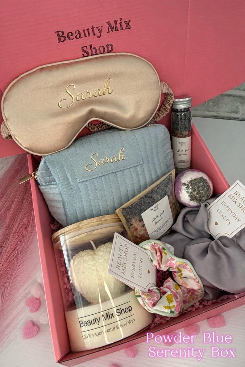 Op de afbeelding: Een roze geschenkdoos van "Beauty Mix Shop" gevuld met zelfzorgartikelen. Bevat een gouden satijnen oogmasker en een poederblauwe make-uptas, beide geborduurd met "Sarah". Bevat ook een kaars, badzout en een scrunchie.