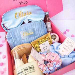 Op de afbeelding: Een roze geschenkdoos met de tekst "Sky Dream Box" gevuld met zelfzorgartikelen. Inclusief een lichtblauw slaapmasker en cosmetische tas geborduurd met "Olivia", een kaars, badzout en een scrunchie. De doos is van "Beauty Mix Shop".