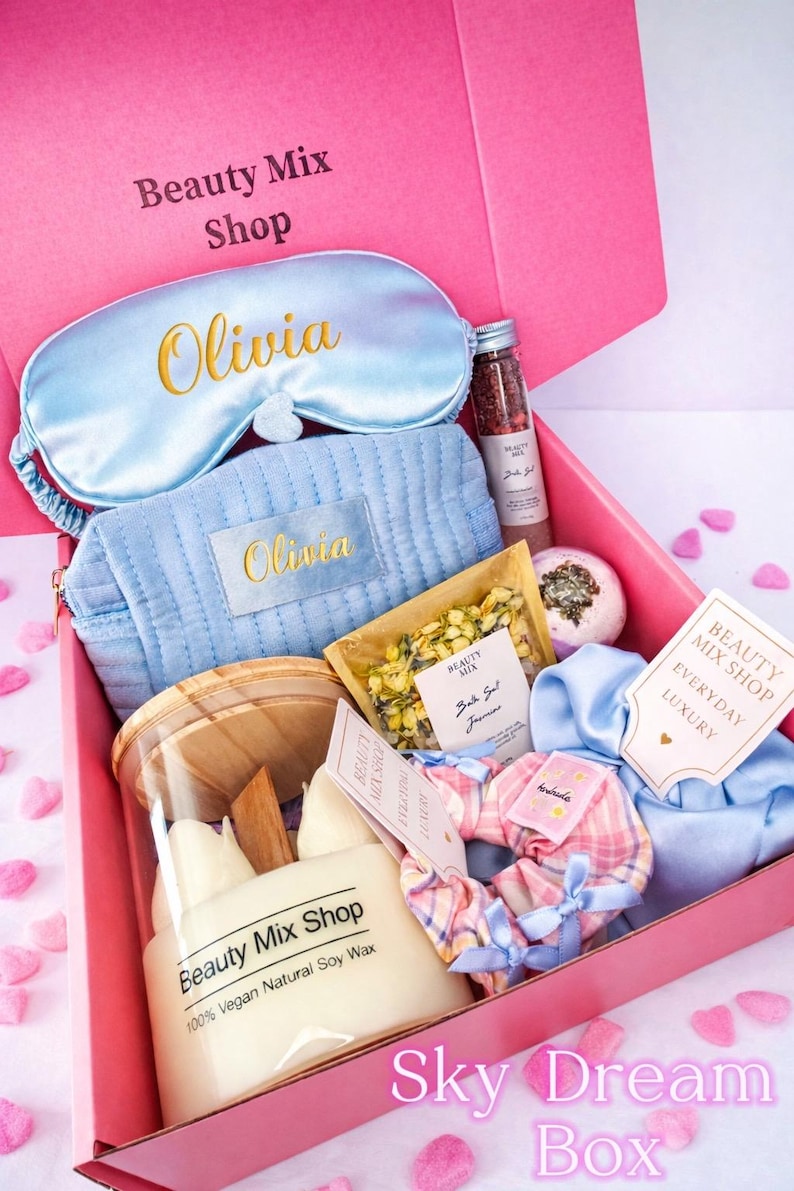 Op de afbeelding: Een roze geschenkdoos met de tekst "Sky Dream Box" gevuld met zelfzorgartikelen. Inclusief een lichtblauw slaapmasker en cosmetische tas geborduurd met "Olivia", een kaars, badzout en een scrunchie. De doos is van "Beauty Mix Shop".