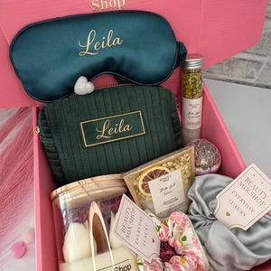 Op de afbeelding: Een open roze geschenkdoos gevuld met schoonheidsproducten. Inclusief een teal oogmasker en cosmetische tas, beide met "Leila" in gouden letters. Bevat ook een kaars, badzout en een bloemen scrunchie. De doos is gelabeld met "Beauty Mix Shop" en "Emerald Calm Box."