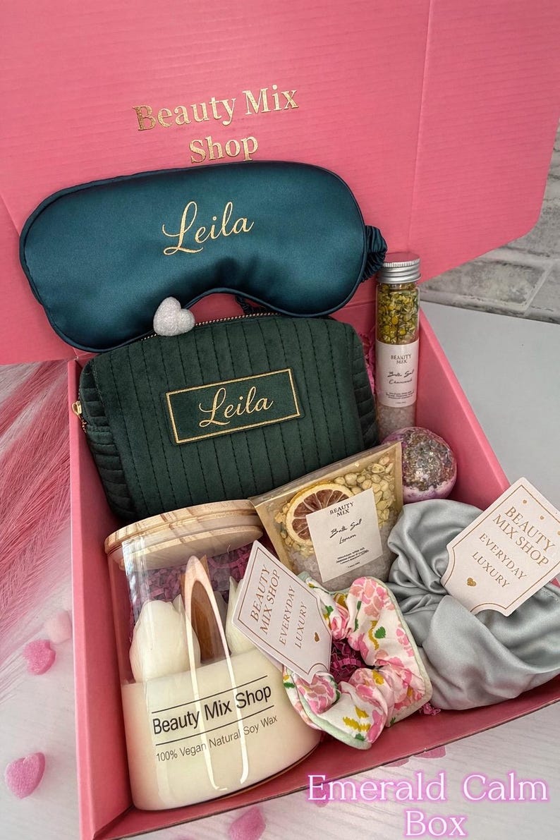 Op de afbeelding: Een open roze geschenkdoos gevuld met schoonheidsproducten. Inclusief een teal oogmasker en cosmetische tas, beide met "Leila" in gouden letters. Bevat ook een kaars, badzout en een bloemen scrunchie. De doos is gelabeld met "Beauty Mix Shop" en "Emerald Calm Box."