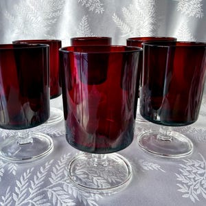 May include: Six ruby red stemmed glasses. The glasses have a cylindrical shape with a clear, circular base. The rich colour of the glass is visible throughout the body. The glasses are arranged on a white tablecloth.