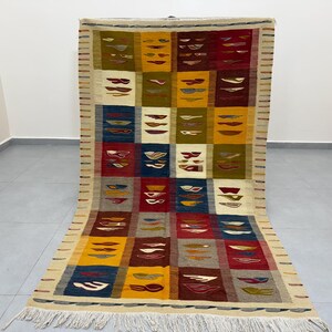 Peut inclure: Un tapis rectangulaire tissé à la main avec un motif géométrique. Le tapis présente une grille de carrés colorés dans les tons de rouge, jaune, bleu et vert, chacun contenant des formes abstraites. Le tapis a une frange crème sur le bord inférieur.