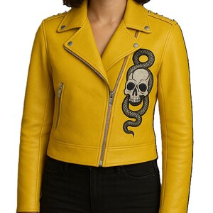 May include: Mustard yellow leather jacket featuring a skull and snake motif. The biker-style jacket has a zip closure, notched lapels, and silver studs on the shoulders. The jacket is made of leather.