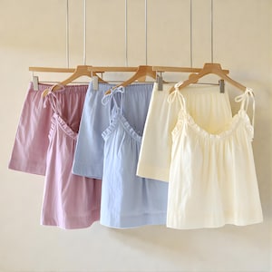 May include: Four sets of pastel-colored pajama sets hanging on wooden hangers. Each set includes a camisole top with ruffled trim and shorts. Colors include pink, light blue, and cream. The camisoles have tie straps.