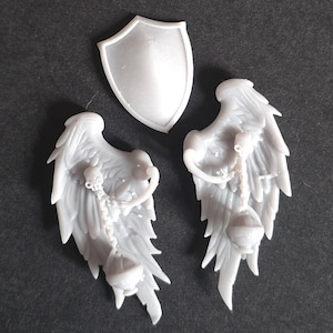 May include: A set of three unpainted gray resin miniature components consisting of a smooth, pointed shield and two detailed feathered wings. Each wing features a sculpted chain hanging from the upper section, ending in a small, rounded censer.