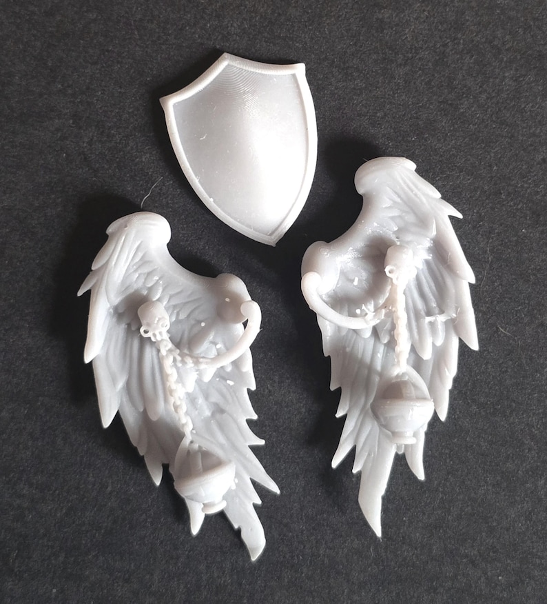 May include: A set of three unpainted gray resin miniature components consisting of a smooth, pointed shield and two detailed feathered wings. Each wing features a sculpted chain hanging from the upper section, ending in a small, rounded censer.