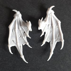May include: Two light gray, molded plastic bat-like wings featuring a skeletal structure with elongated, pointed finger-like supports and thin, textured membranes between the bones. Each wing has a sharp, curved claw at the top apex.