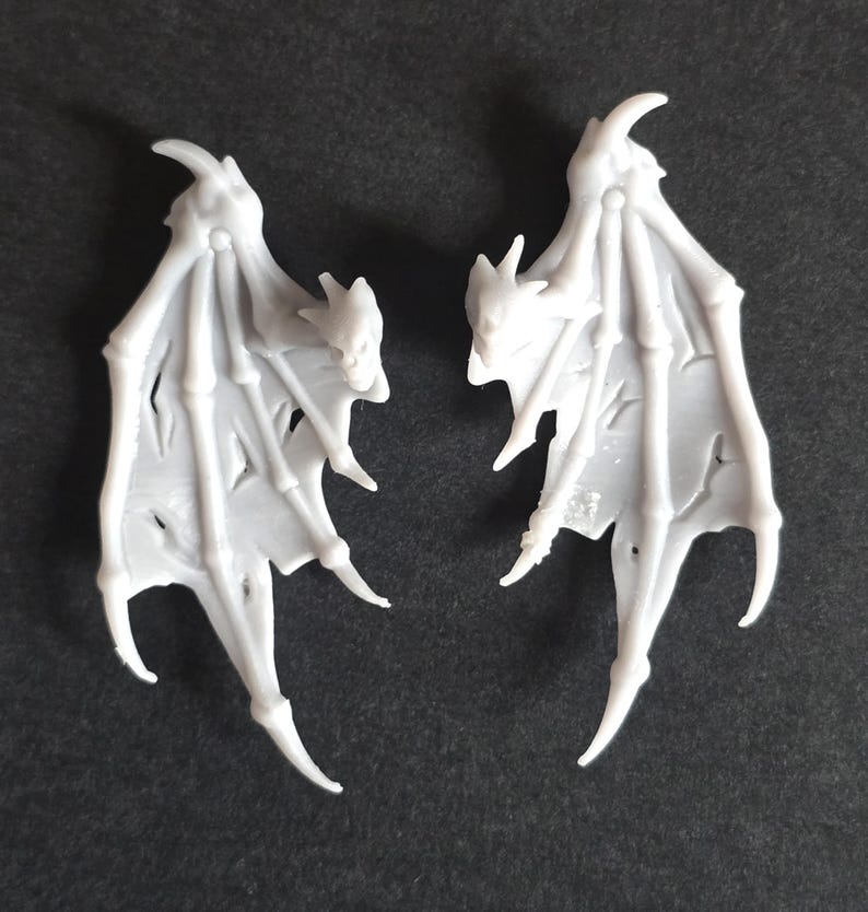May include: Two light gray, molded plastic bat-like wings featuring a skeletal structure with elongated, pointed finger-like supports and thin, textured membranes between the bones. Each wing has a sharp, curved claw at the top apex.
