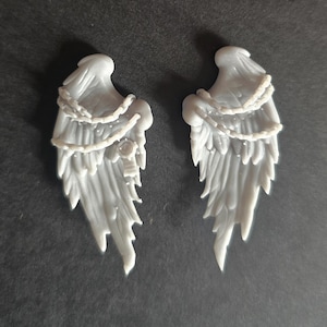 May include: A pair of light gray, molded resin angel wings featuring detailed feather textures and a draped chain element across the upper section. The left wing includes a small circular emblem detail, while both pieces exhibit a smooth, matte finish.