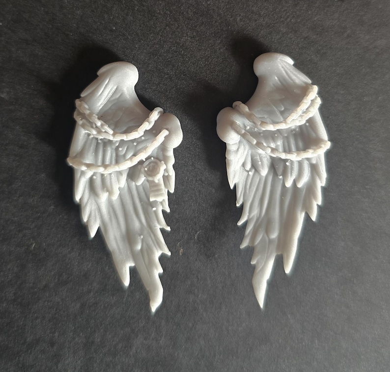 May include: A pair of light gray, molded resin angel wings featuring detailed feather textures and a draped chain element across the upper section. The left wing includes a small circular emblem detail, while both pieces exhibit a smooth, matte finish.