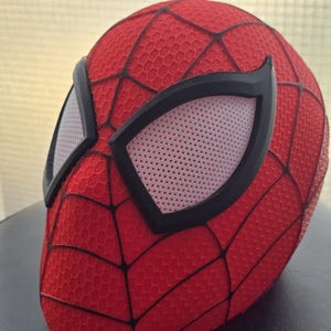 Pode incluir: Uma máscara vermelha do Homem-Aranha com uma superfície texturizada e detalhes de teia de aranha pretos. As aberturas dos olhos são brancas com um padrão de malha, emolduradas por bordas pretas. A máscara é uma réplica do icônico capacete do super-herói.
