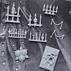 Op de afbeelding: Een verzameling grijze hars miniatuur spelonderdelen, waaronder vier heksecties met punten en schedels, twee hangende kettingaccessoires, een stervormig embleem met drie schedels en een rechthoekige plaat met een gezichtontwerp.