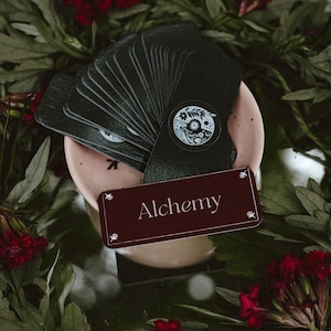 May include: A deck of black tarot cards with a white and black circular design on the back, fanned out on a light pink plate. A burgundy card with the word "Alchemy" is in front of the deck. The cards are surrounded by green leaves and red flowers.