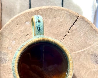 May include: A ceramic mug filled with a dark brown liquid, possibly coffee or tea. The mug has a teal and brown glaze and a rounded handle. It sits on a wooden surface, creating a rustic aesthetic.