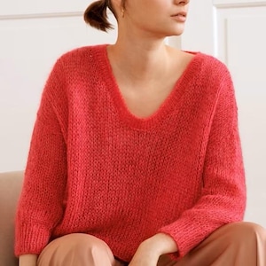 May include: A coral-colored, V-neck sweater with long sleeves. The sweater has a loose knit texture and is paired with light brown pants. The model is sitting down, with a neutral background.