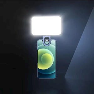 May include: A white rectangular LED light attached to a smartphone with a clip. The phone has a green and yellow gradient background. The light is designed to illuminate the phone's camera.