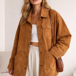 Könnte beinhalten: Braune Wildlederjacke mit Kragen, Knopfverschlüssen und zwei Vordertaschen. Die Jacke wird über einem weißen Oberteil und mit cremefarbenen Hosen getragen. Eine braune Leder-Umhängetasche ist ebenfalls zu sehen.
