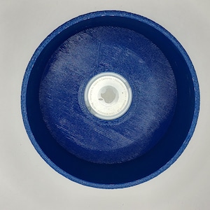 Puede incluir: Vista superior de un recipiente circular azul oscuro con una superficie texturizada. En el interior, un objeto circular blanco y plateado está centrado. El borde del recipiente tiene un patrón tejido. El fondo es una superficie blanca lisa.