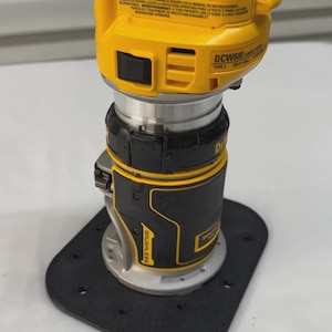 May include: A yellow and black cordless router with the text "DCW600" and "BRUSHLESS". The tool has a silver base and sits on a black base plate. The top of the router has safety warnings.