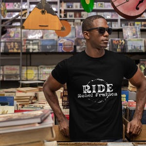 May include: Black t-shirt with the text "RIDE Rebel Fruitica" in a white, distressed font. The shirt is worn by a person wearing sunglasses, standing in a vintage shop filled with books and records.