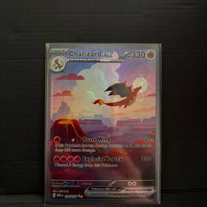 May include: A Charizard EX Pokémon card featuring a flying orange dragon with black wings against a sunset landscape. The card includes the text "Charizard EX", "Brave Wing", and "Explosive Vortex". The card is in a protective sleeve.