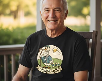 May include: A black t-shirt featuring a graphic of a goose with a white beard and a blue plaid vest, holding a wooden staff. The goose is followed by three small goslings in a grassy field under a sun and clouds. The text reads GRANDPA GOOSE.