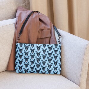 Könnte beinhalten: Hellblaue Umhängetasche mit einem sich wiederholenden Muster aus schwarzen Hundegesichtern. Die Tasche hat einen schwarzen Riemen und Reißverschluss und liegt auf einem beigen Stuhl. Ein brauner Blazer ist über den Stuhl drapiert.