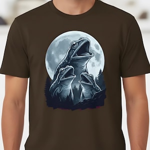 May include: Brown t-shirt featuring a detailed illustration of three frogs singing under a full moon. The frogs are in shades of blue and gray, with a forest silhouette in the background. The design is centered on the shirt.