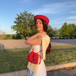 May include: A bright red crocheted beret and a two-toned drawstring bag featuring a red crocheted top section and a cream-coloured crocheted bottom section, worn with a white dress and a cream-coloured sweater draped over the shoulders.