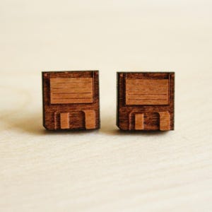 Puede incluir: Un par de pendientes de madera con forma de disquetes vintage. Los pendientes son de color marrón oscuro con un detalle marrón más claro en la parte delantera.